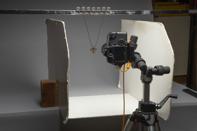 Camera on tripod aimed at necklace hanging off a crossbar; two white poster board pieces on each side to bounce light