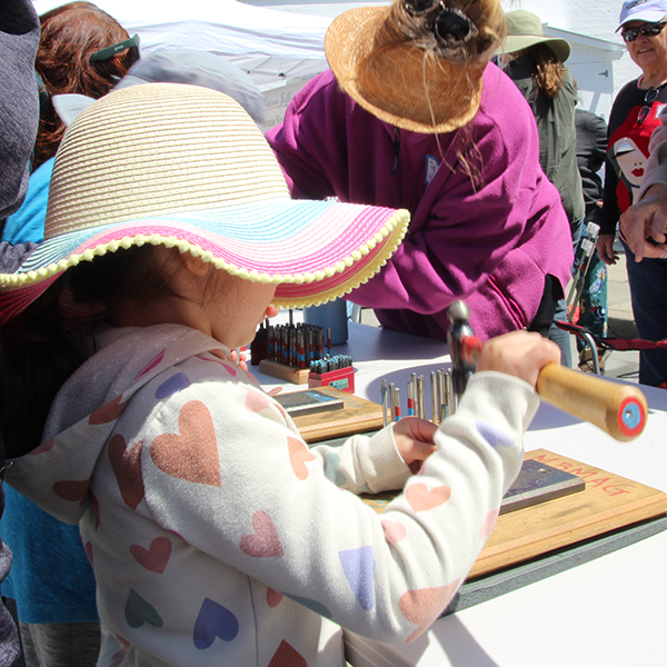 A young participant stamps a shape into a copper strip to create a bracelet at the Make A Bracelet event as part of the Monterey Museum of Art Block Party 2023