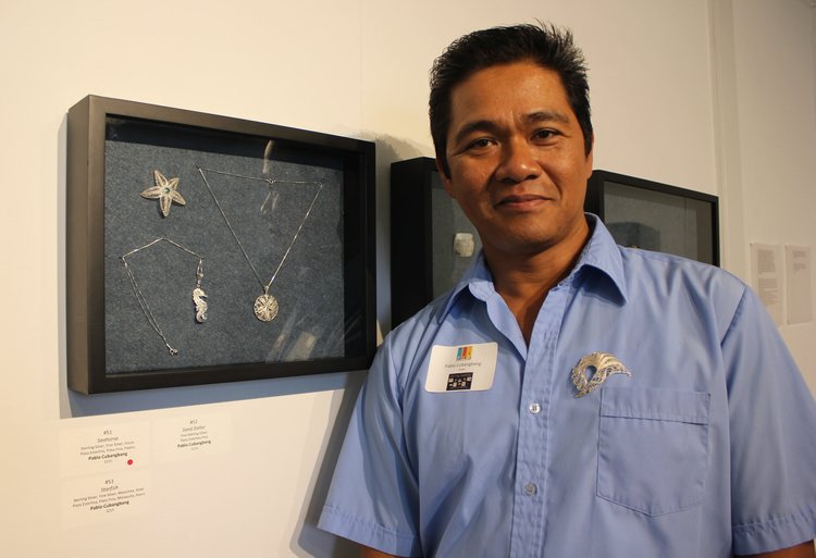 Portrait of Pablo Cubangbang standing next to a wall case containing 3 of his filigree jewelry pieces. A star shape, a round pendant necklace, and a seahorse necklace.